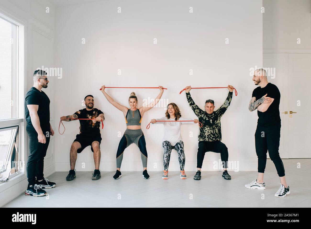 Istruttori Fitness osservando amici facendo squat contro la parete con banda di resistenza in studio Foto Stock