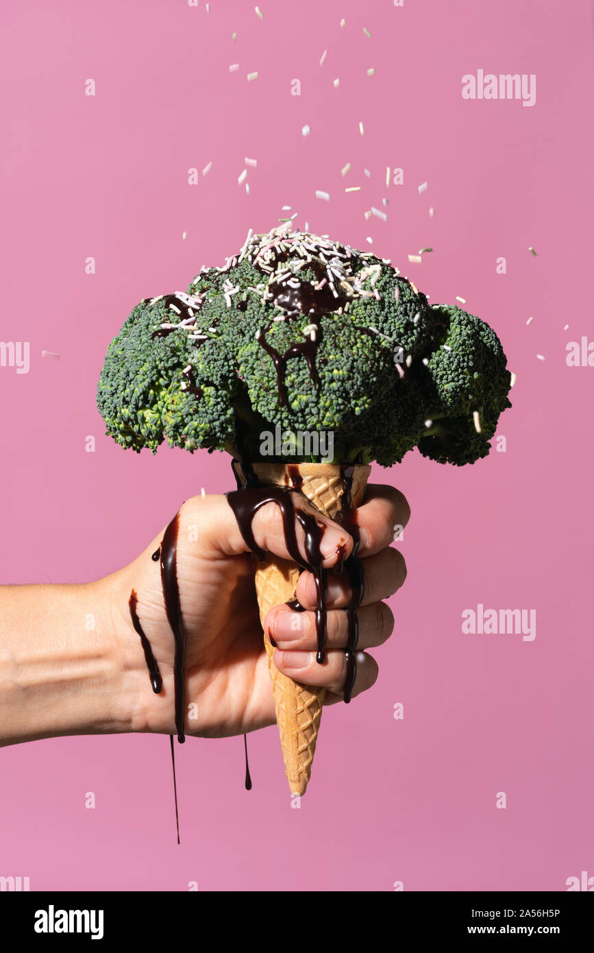 Studio colpo di mano d'uomo tenendo cono gelato con broccoli in cima, gocciolamento in salsa di cioccolato e spruzza, contro sfondo rosa Foto Stock