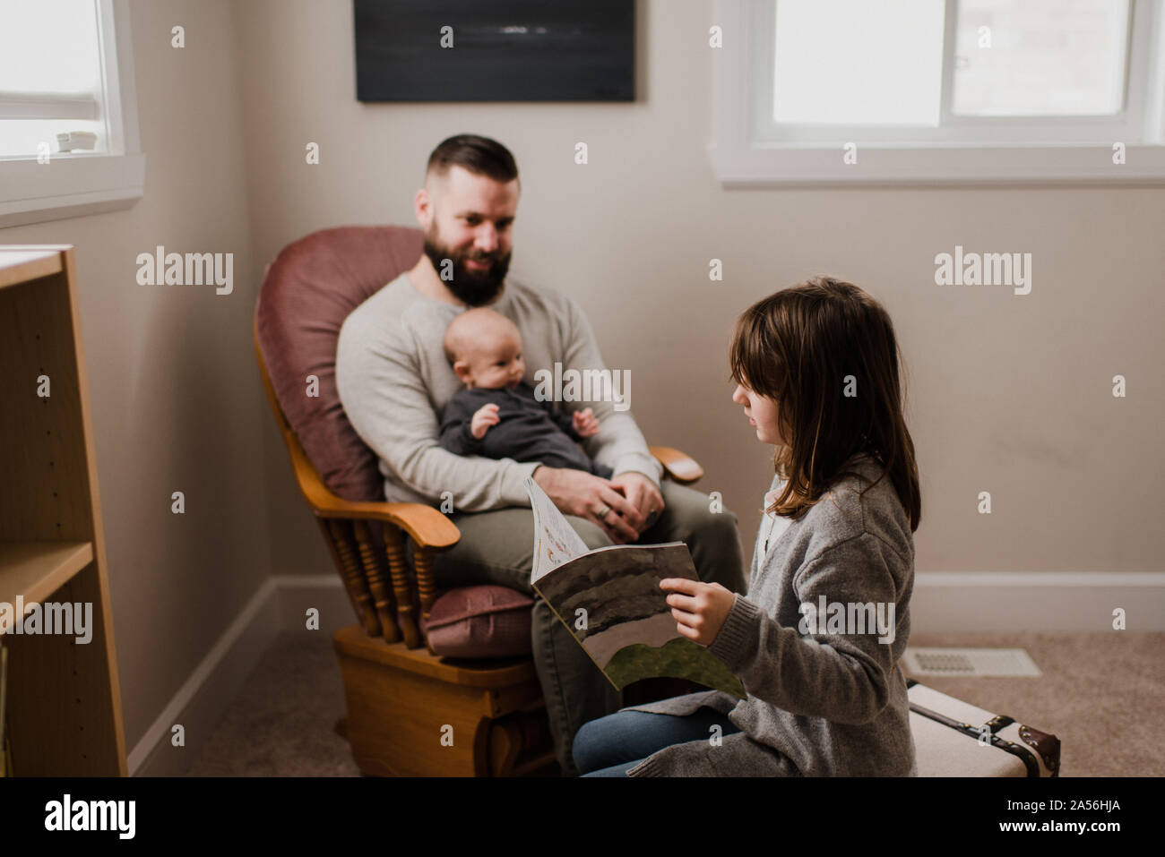 Ragazza nel soggiorno leggendo il libro vicino al padre e fratello di bambino su poltrona Foto Stock