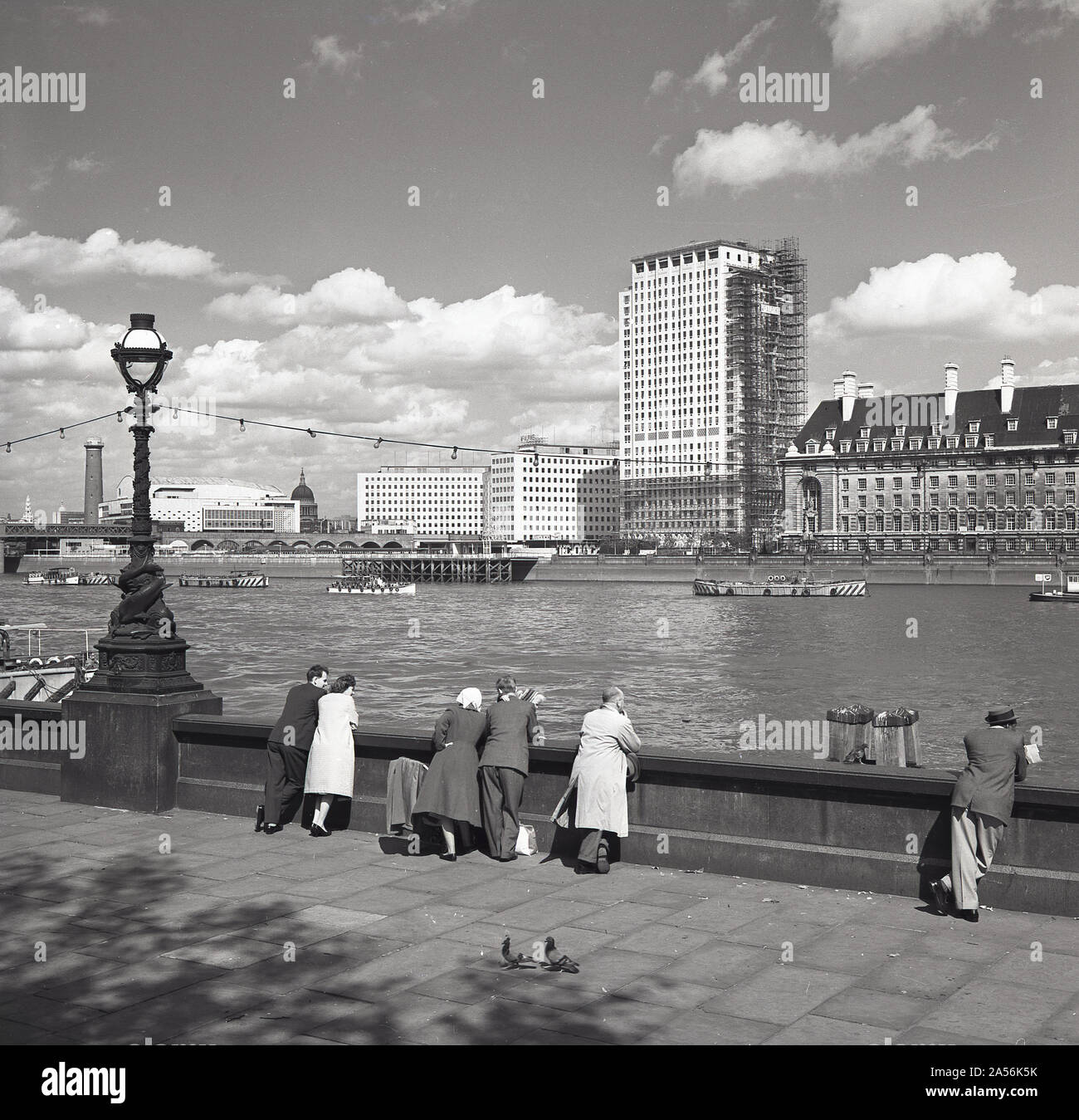 1960, storico, persone in piedi sul terrapieno victoria appoggiate sulla barriera di marciapiede che guarda sul Tamigi, verso Southbank, Londra, Inghilterra, Regno Unito. Sul lato sud del fiume, la County Hall, sede del GLC dal 1965 al 1986, un nuovo e moderno edificio a torre in costruzione e, all'estrema sinistra, la Shot Tower accanto alla Royal Festival Hall. Foto Stock