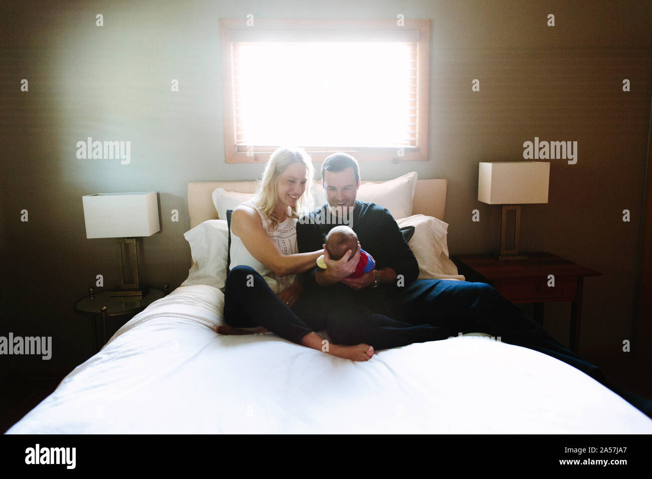 Due nuovi genitori sorriso al loro figlio neonato mentre è seduto su un letto Foto Stock