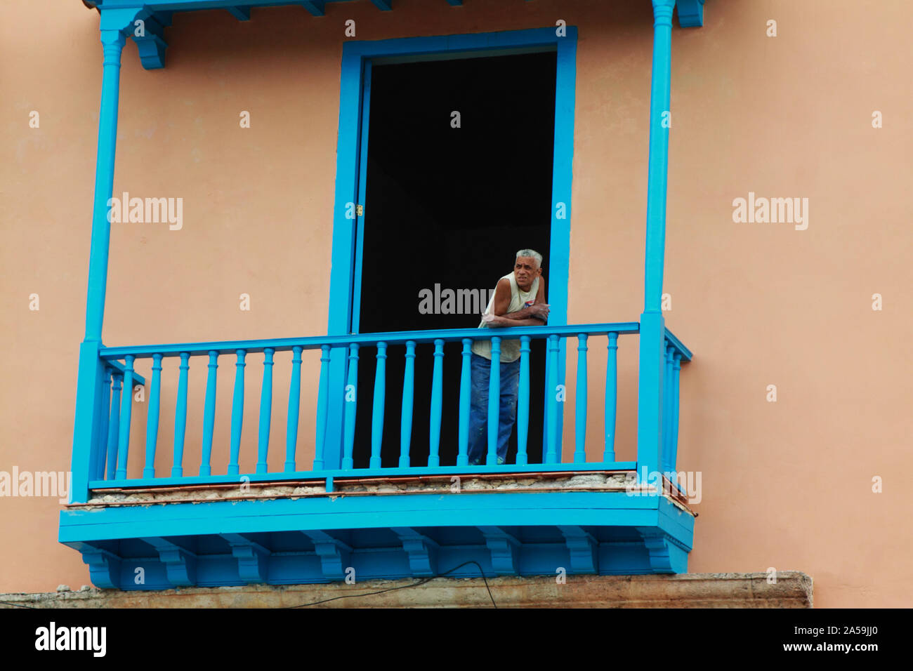 Uomo cubano appoggiata contro il suo blu ringhiera di balcone e visualizzazione della vita di strada sotto. Foto Stock