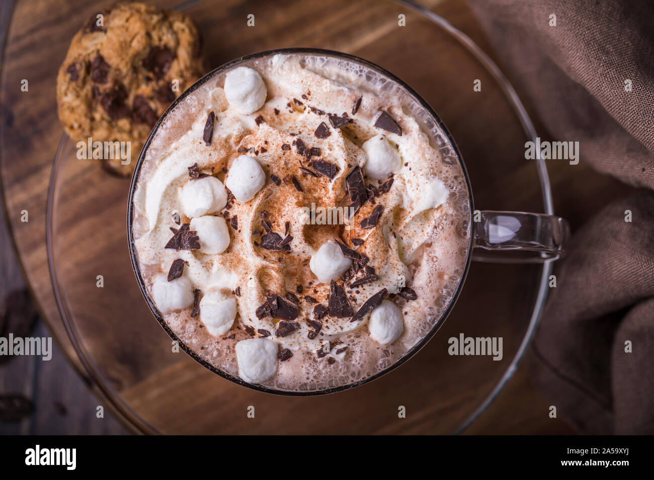 Una tazza di cioccolata calda con panna montata, marshmallows e cacao visto da sopra. La coppa è su un coperchio di vetro con un Chocolate Chip Cookie sul lato. Foto Stock Una tazza di cioccolata calda con panna montata, marshmallows e cacao visto da sopra. La coppa è su un coperchio di vetro con un Chocolate Chip Cookie sul lato. Foto Stock