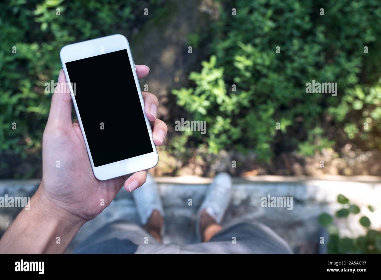 Mockup immagine di una mano di un uomo bianco di contenimento mobile phone con schermata nera vuota mentre si sta in piedi sul calcestruzzo pavimento Lucidatura con verde lasciare e natura Foto Stock