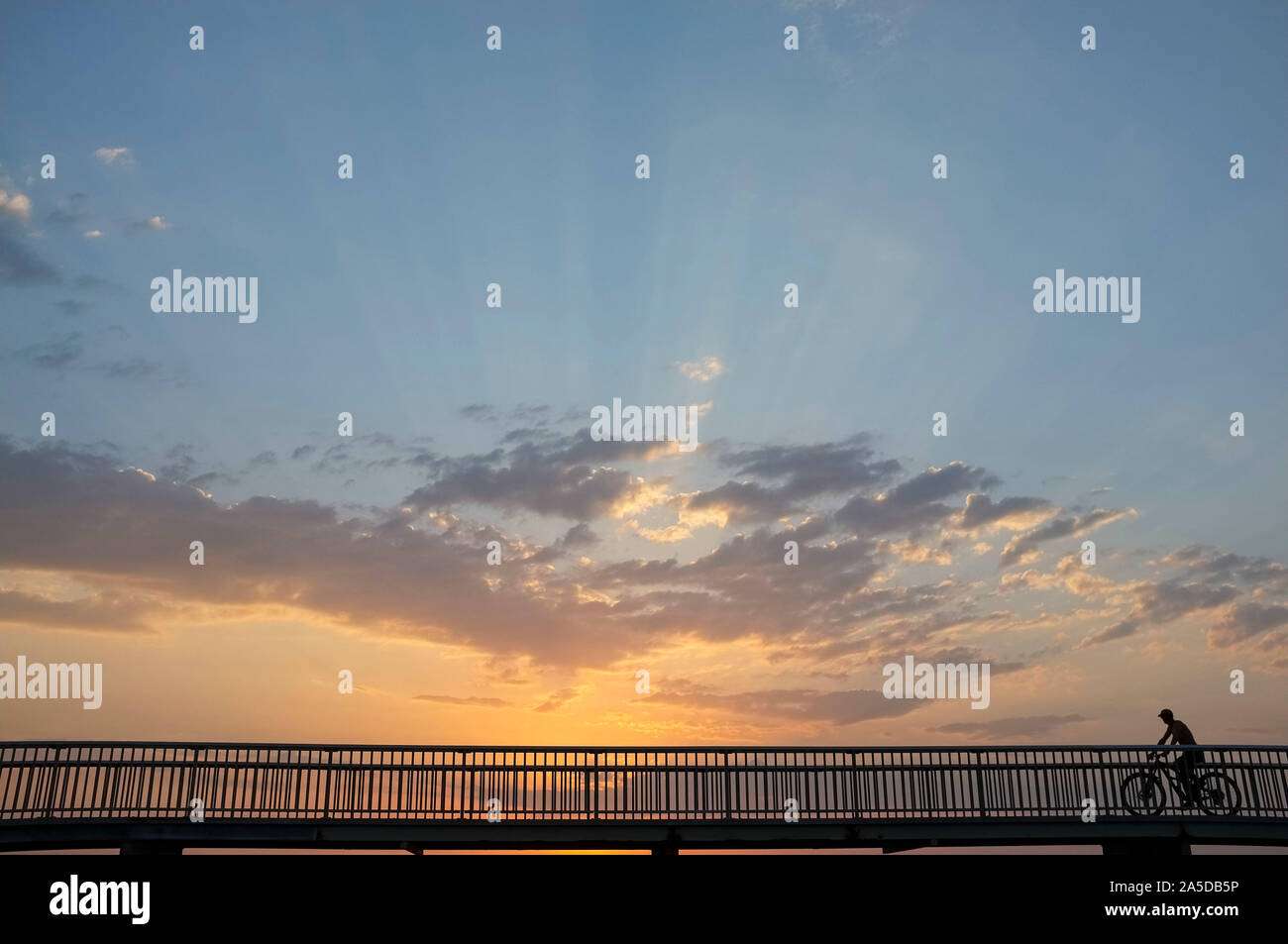 Silhouette di un uomo in sella ad una bicicletta su un piccolo ponte, contro un tramonto. Foto Stock