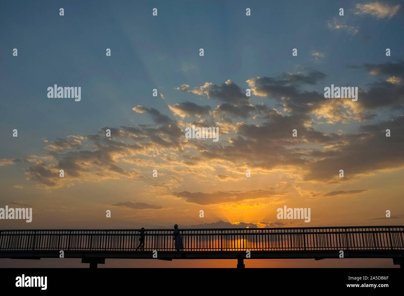 La silhouette di una donna e di un bambino che cammina verso ogni altro su un ponte pedonale al tramonto. Foto Stock