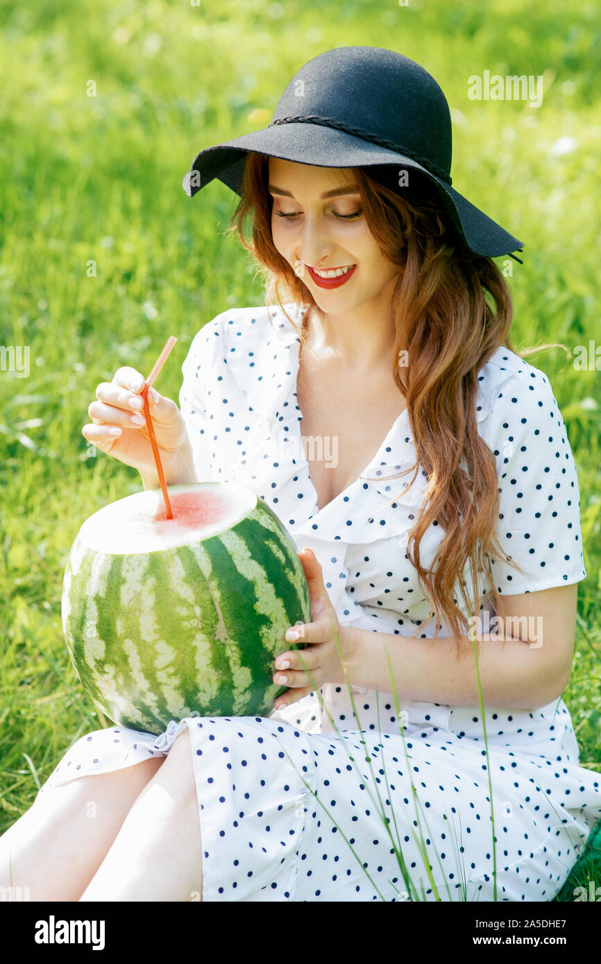 Pretty Woman in Black Hat detiene un intero anguria con cocktail di paglia. Ritratto di ragazza sorridente in black hat con un cocomero intero in natura. Foto Stock