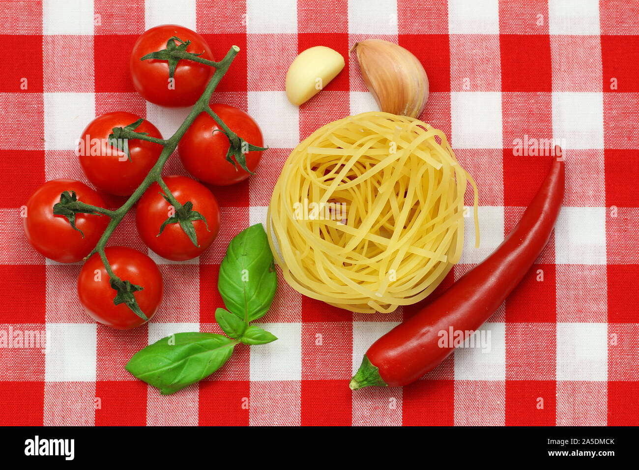 Materie prime: spaghetti, pomodori ciliegini, peperoncino rosso, spicchi di aglio e di foglie di basilico fresco su rosso e bianco di panno a scacchi Foto Stock