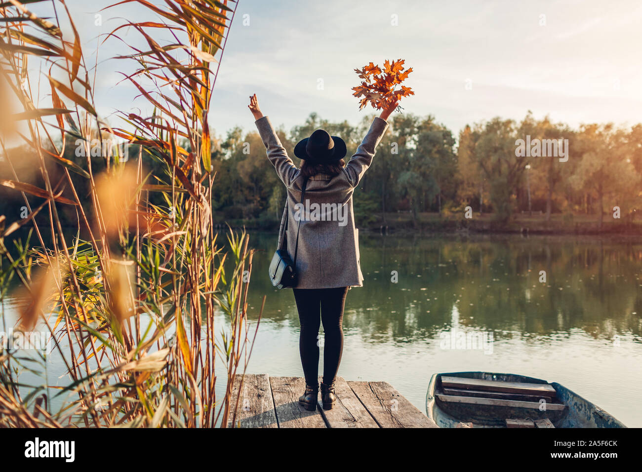 Donna felice camminando sul molo sul lago in barca alzando le mani e ammirando il paesaggio autunnale. Caduta stagione attività Foto Stock