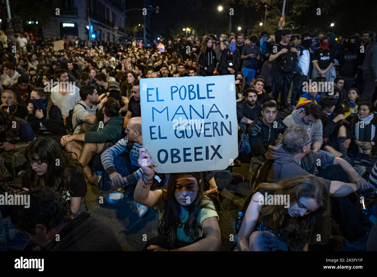 Un manifestante detiene una targhetta che dice la gente regola il governo obbedisce accompagnati da numerosi manifestanti seduti di fronte al Via Laietana stazione della polizia durante la manifestazione.il sesto giorno di protesta dopo l'annuncio delle sentenze da parte della Corte suprema di Spagna che condanna il catalano e i leader politici a lunghe pene detentive. Il agenti antisommossa hanno ripetutamente caricato i manifestanti intorno della stazione centrale di polizia della polizia nazionale in Via Laietana. Foto Stock