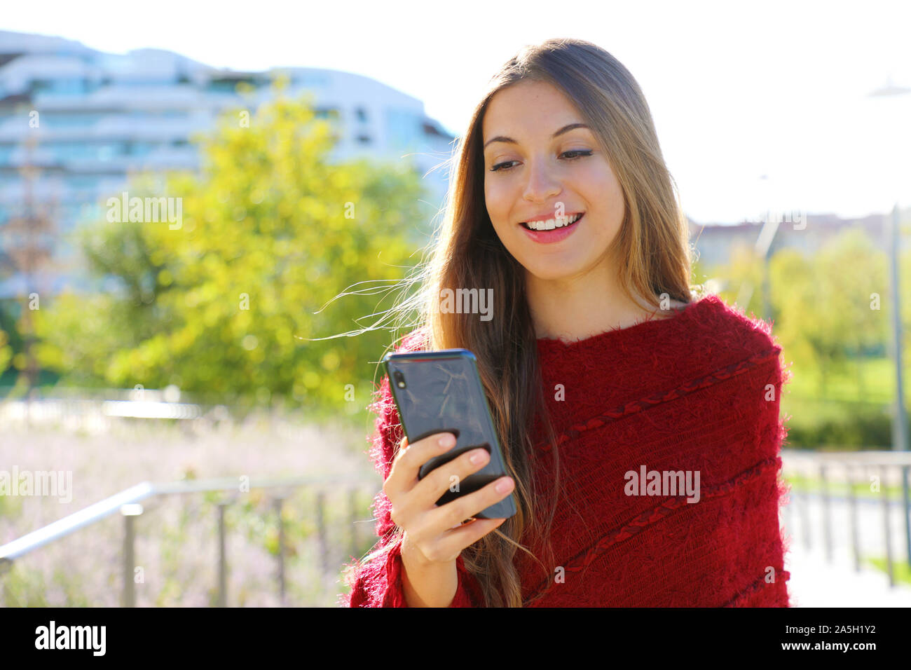 Ragazza texting su smart phone nel parco della città indossando un poncho rosso. Foto Stock