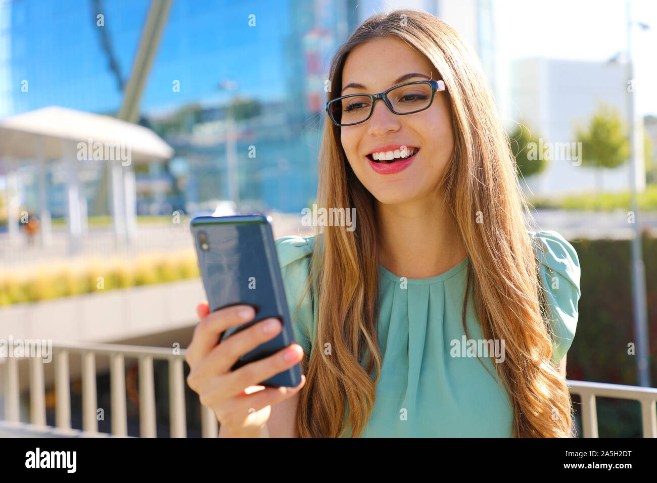 Sorridente smart business donna con telefono cellulare in strada con edifici per uffici in background. Foto Stock