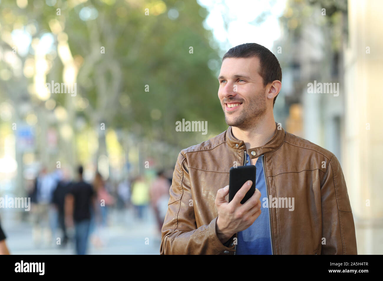 Vista frontale - Ritratto di un uomo felice azienda smart phone camminando verso la telecamera guardando al lato della strada Foto Stock