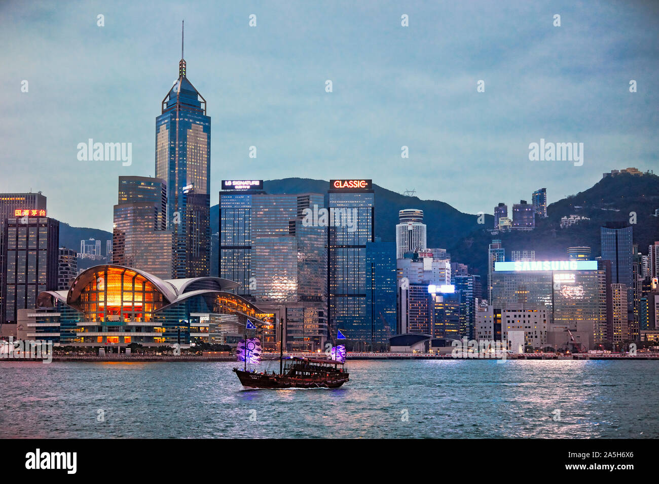 Tradizionale giunca nel Victoria Harbour passando da edifici del Central Waterfront illuminati al tramonto. Hong Kong, Cina. Foto Stock
