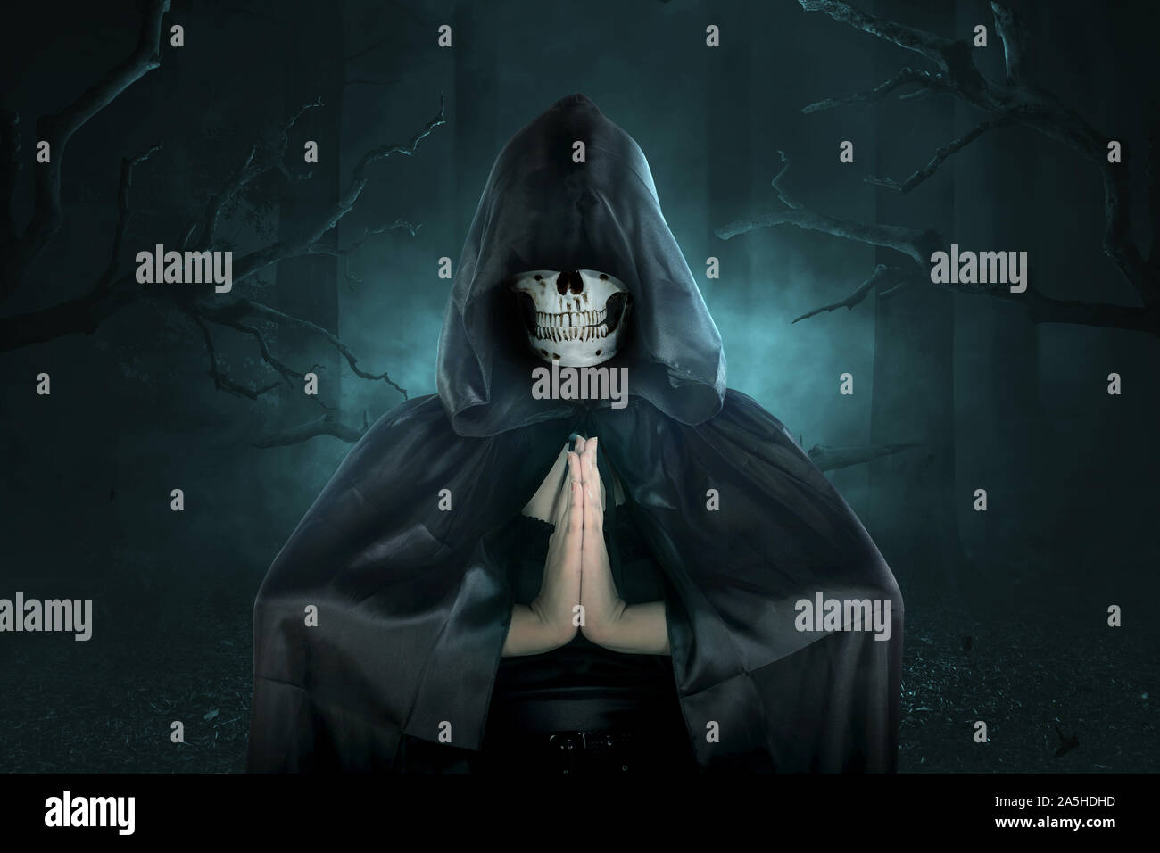 Strega con teschio umano in testa con cappuccio nero facendo magia rituale sulla foresta scura Foto Stock