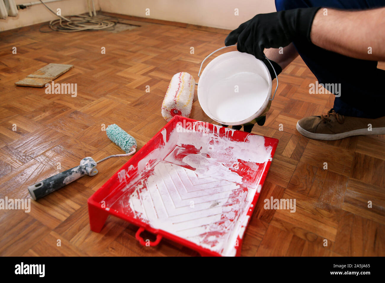 Operaio edile finitura facendo lavori di rinnovo nell'appartamento. Pittore con guanti neri si riversa il colore bianco dalla benna nella vaschetta di colore rosso per la miscelazione della vernice. Foto Stock