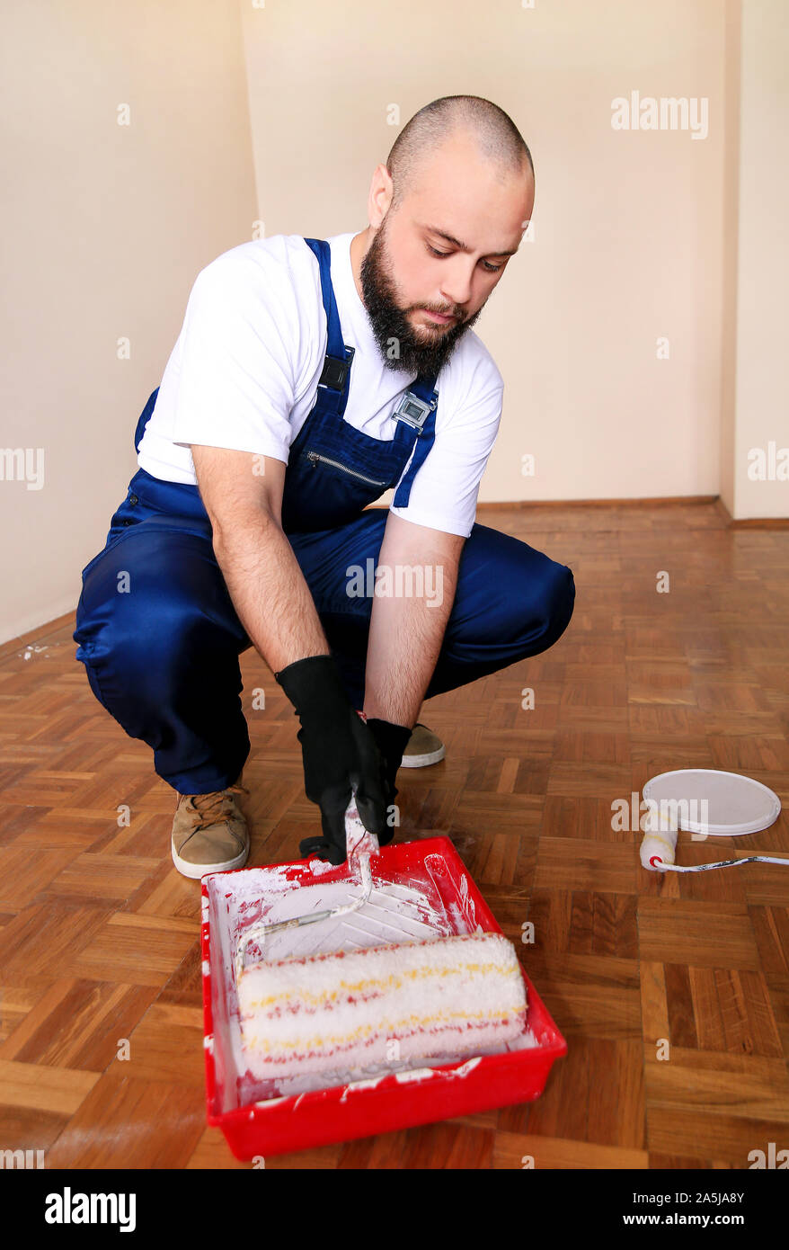 Operaio edile finitura facendo lavori di rinnovo nell'appartamento. Pittore con guanti bianchi mantiene la spazzola a rullo in mano e si mescola la vernice nel contenitore. Foto Stock
