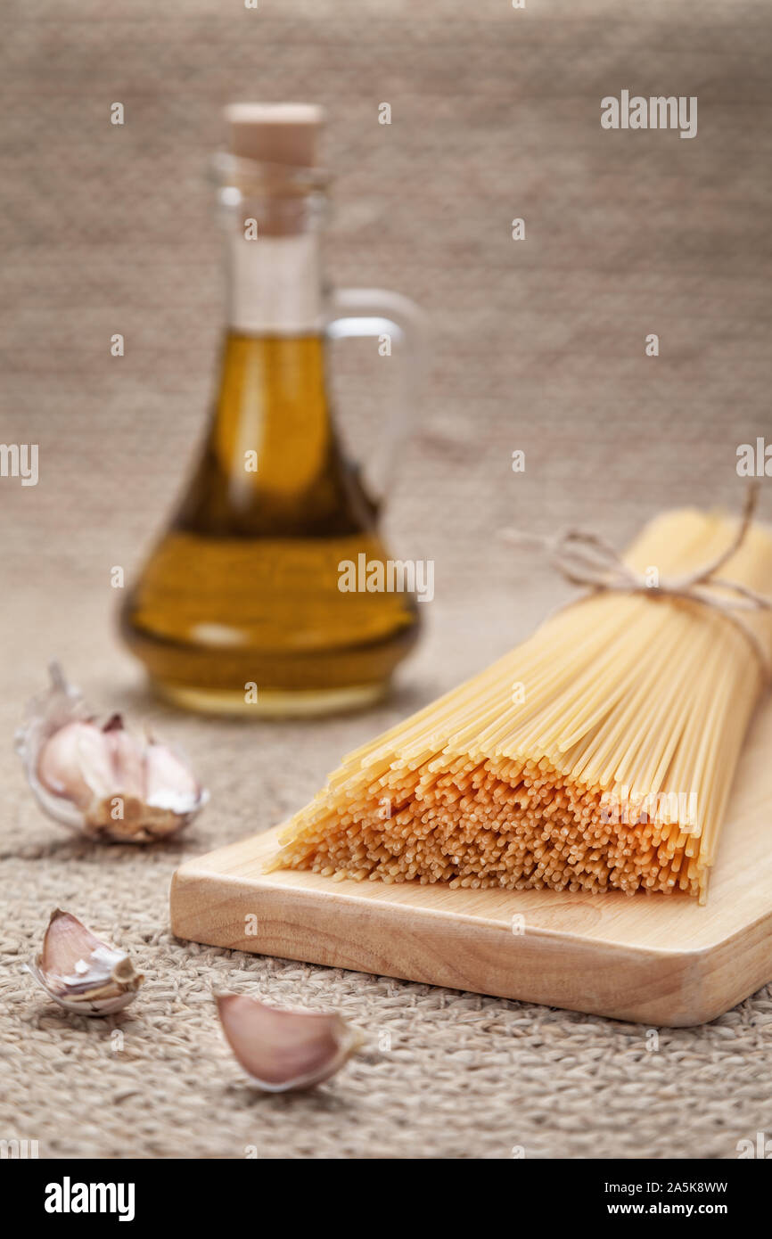 Olio di cottura e ingredienti. Spaghetti legati in mazzi da corda artigianale giacente sulla superficie naturale bulbi di aglio. Caraffa con olio di oliva sullo sfondo Foto Stock