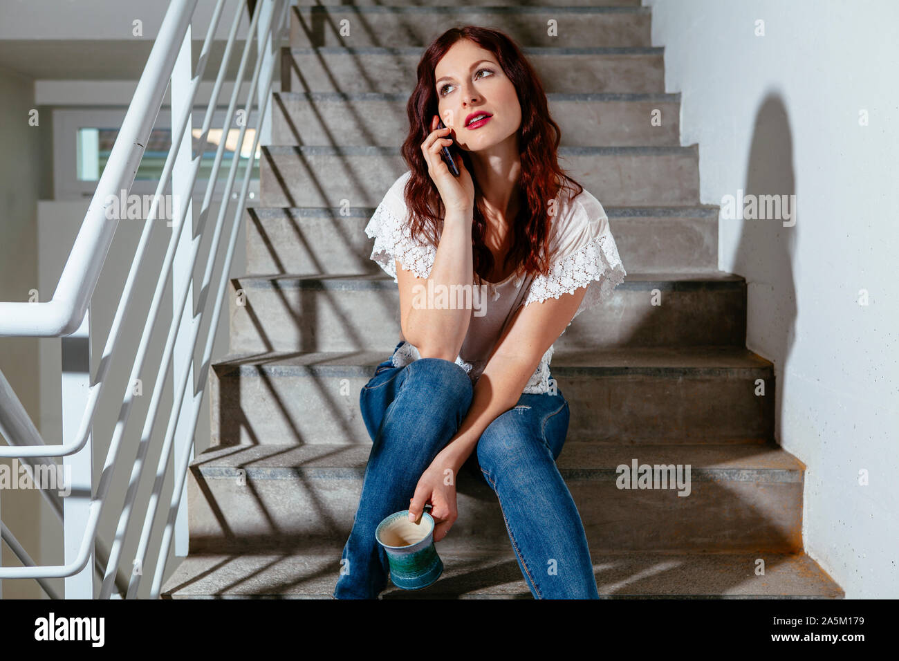 Giovane donna con i capelli rossi seduta sulle scale e parlando sul suo smartphone e di bere il caffè. Foto Stock