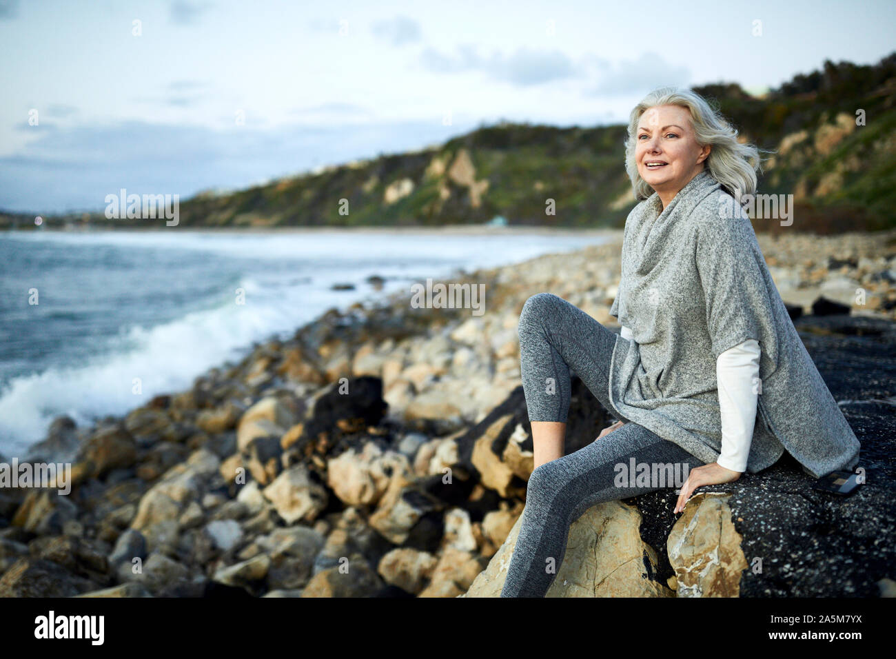 Vista laterale di sorridere senior donna seduta su roccia a Manhattan Beach durante il tramonto Foto Stock