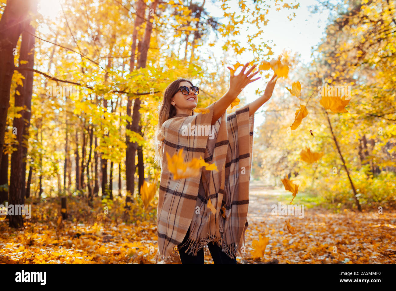 Caduta stagione attività. Donna gettando le foglie in autunno foresta al tramonto. Giovane donna avendo divertimento all'aperto indossare abbigliamento elegante Foto Stock