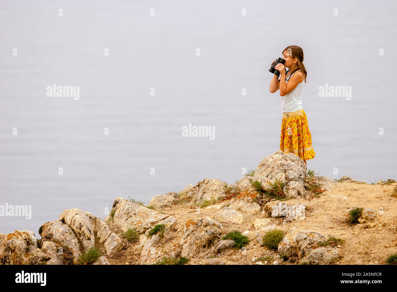 Una ragazza con un binocolo nelle sue mani sorge sul lago Baikal sulle rocce e guarda oltre il binocolo. Grosse pietre. T-shirt e colorato di giallo gonna. Se Foto Stock