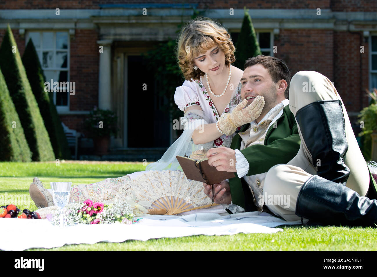 Giovani amanti vestito in abiti d'epoca seduti sulla coperta picnic. Gentleman guardando il suo partner tenendo prenota Foto Stock