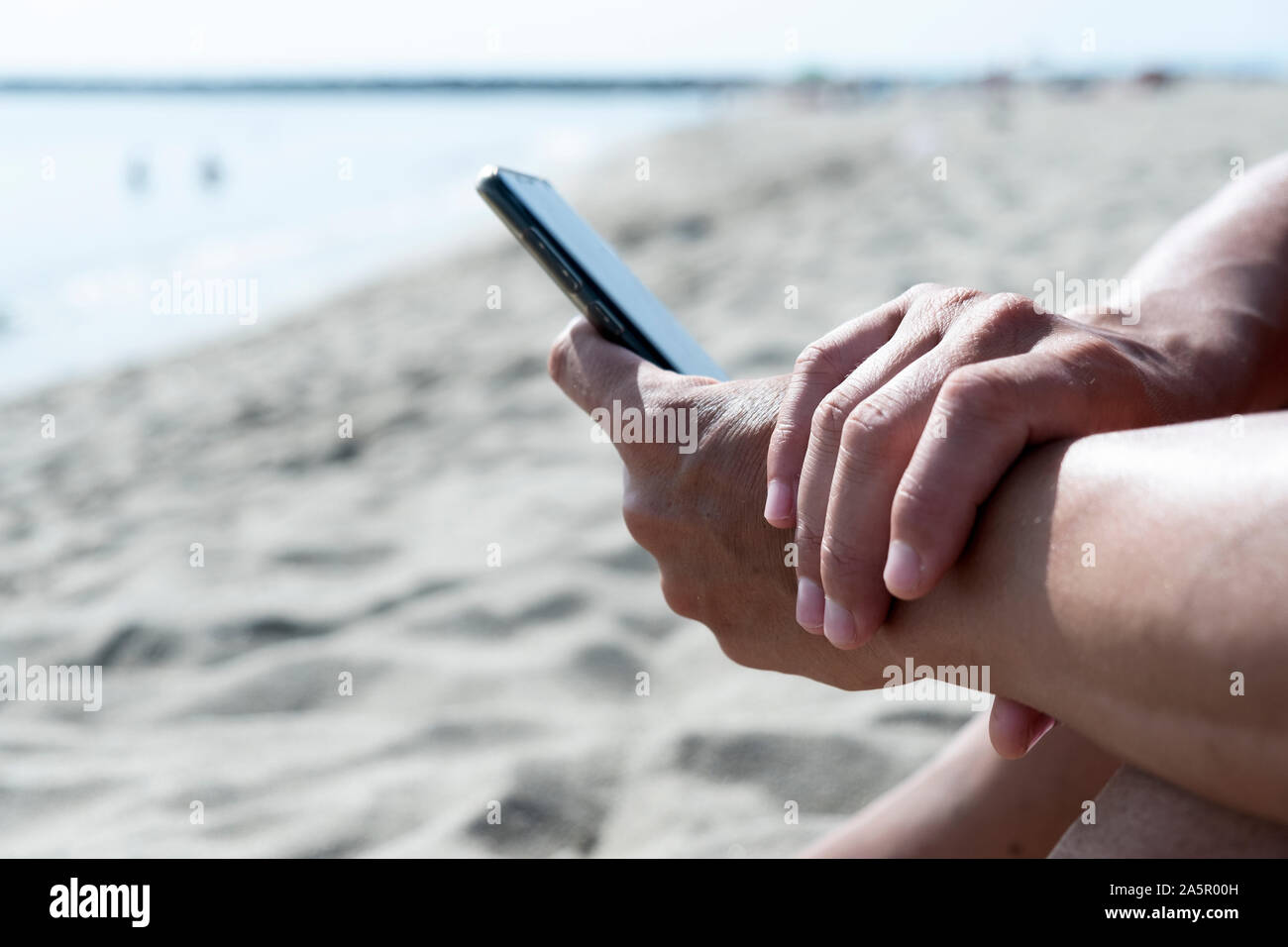Primo piano di un giovane uomo caucasico usando il suo smartphone su di una spiaggia di sabbia bianca accanto al mare Foto Stock