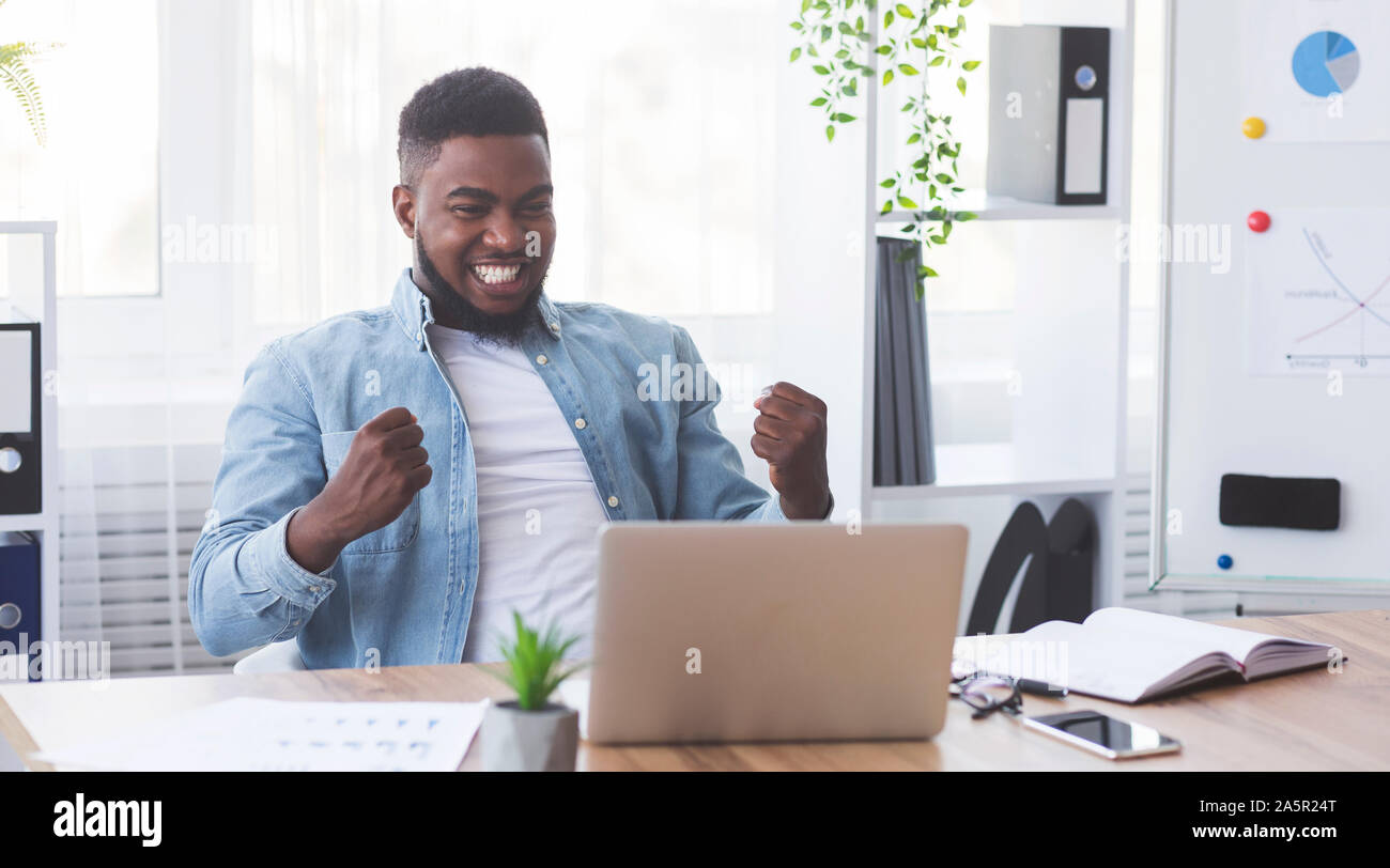 Imprenditore nero guardando laptop, clenching pugni, celebrando il successo aziendale Foto Stock