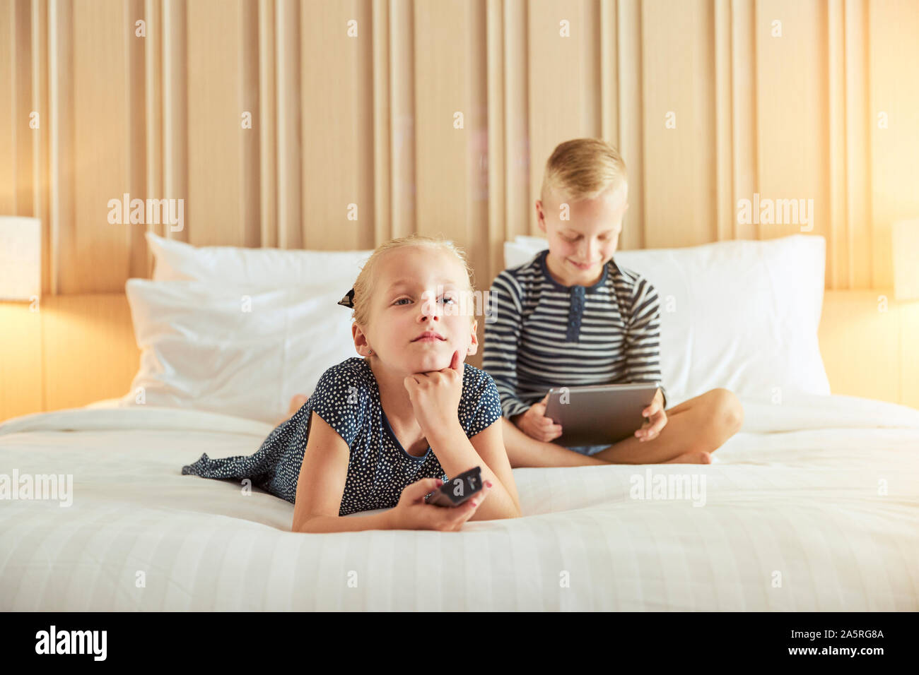 Incantevole piccola ragazza distesa su un letto a guardare la televisione con il suo fratello seduto dietro di lei utilizzando una tavoletta digitale Foto Stock