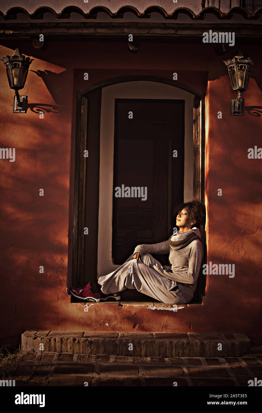 Nero donna seduta sul pavimento in aprire il telaio della porta della sua casa godendo i raggi del sole con gli occhi chiusi Foto Stock