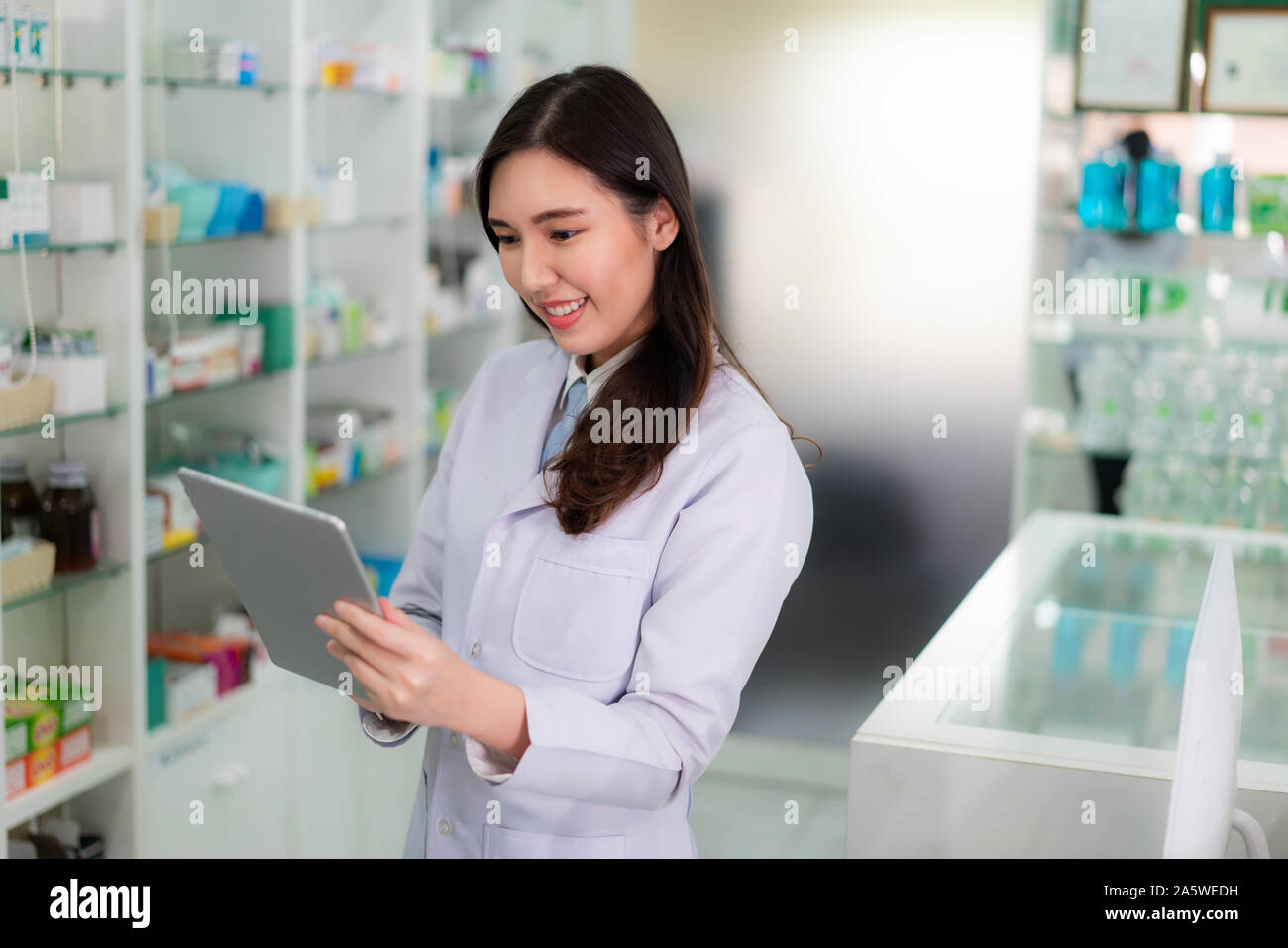 Fiducioso asiatici femmina giovane farmacista con sorriso holding tavoletta digitale e per la ricerca di database prodotto in farmacia farmacia. Medicina, PHA Foto Stock
