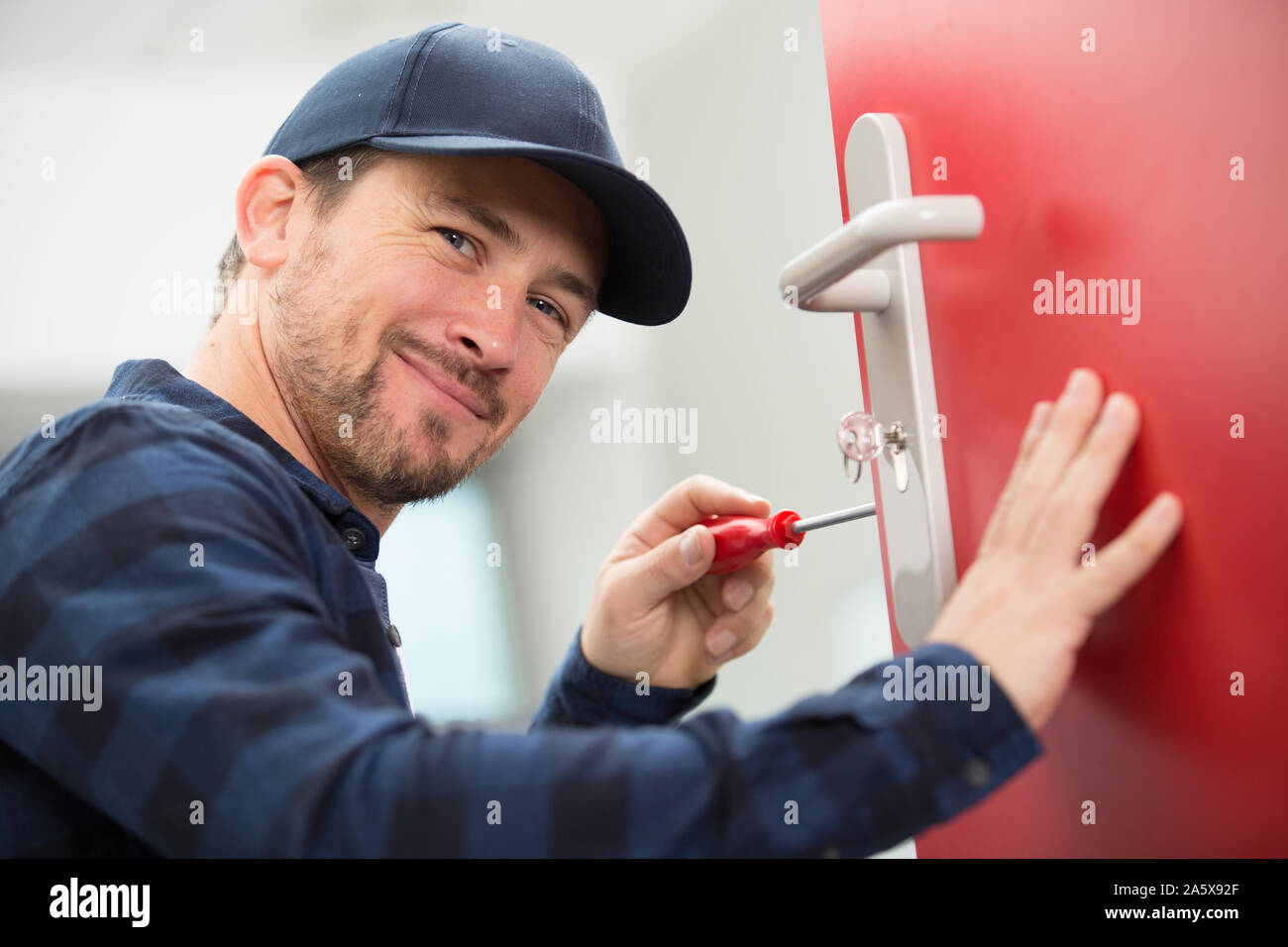 Felice giovane falegname installa serratura della porta con un cacciavite senza fili Foto Stock