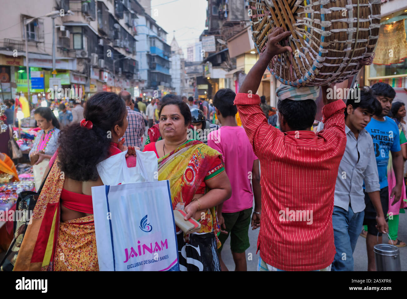 Due indiani casalinghe stop per chattare nella trafficata area di mercato di Bhuleshwar, Mumbai, India, mentre un facchino che porta il suo cesto sulla sua testa passa da Foto Stock