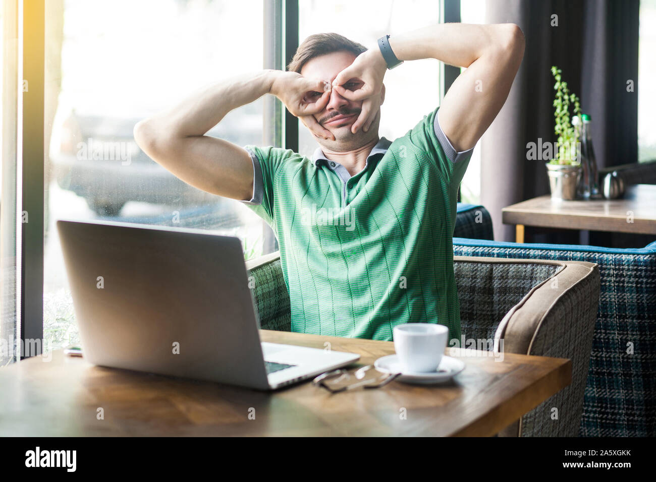 Giovani funny crazy businessman in verde t-shirt seduto e guardando con il binocolo gesto. business e concetto freelancing. interna shot vicino grande vittoria Foto Stock