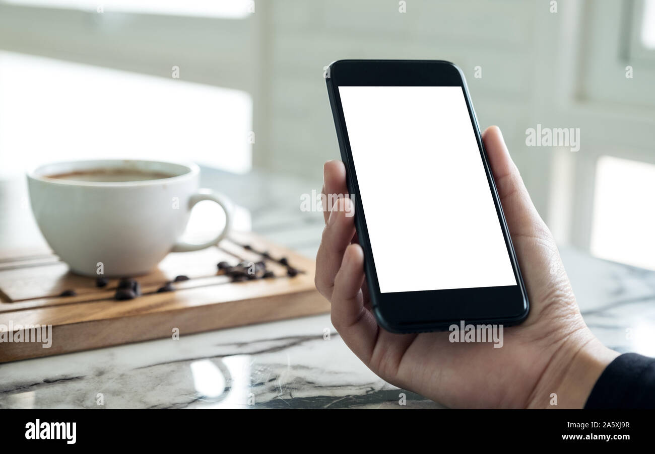 Mockup immagine di mani nero cellulare con schermata bianca vuota con una tazza di caffè nella caffetteria Foto Stock