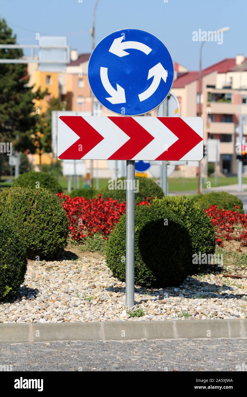 Round blu e bianco rotatoria cartello stradale sopra a strisce rosse e bianche freccia curva pericolosa cartello stradale montata su palo metallico circondato con ghiaia Foto Stock