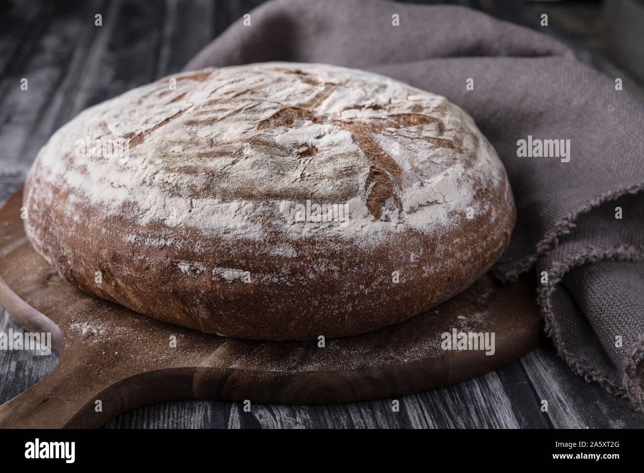 Una casalinga levain rotondo con pane di farina di frumento e segale. che è prevista su tavoli in legno scuro e con un sfondo scuro. Vi è un round tagliere onu Foto Stock