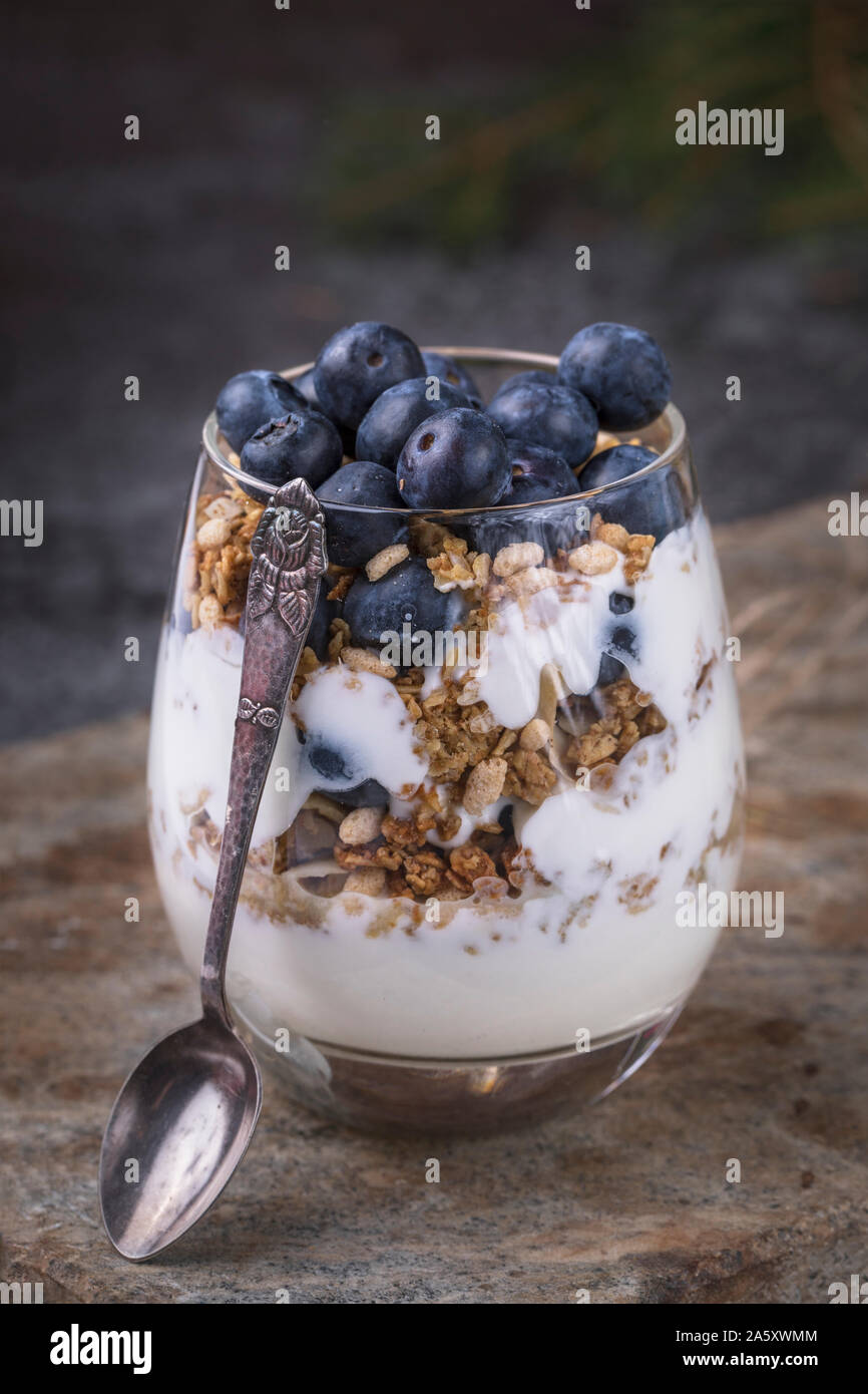 Una fresca e salutare prima colazione di yogurt, cereali fatti in casa e i mirtilli in un bicchiere. Il vetro è su una usura della superficie di pietra e accanto al vetro è un Foto Stock