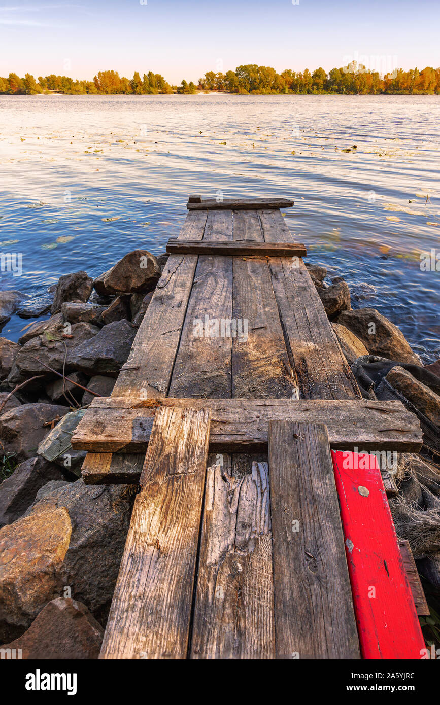 Un pontile in legno con una vernice rossa plank su grosse pietre vicino al blu del fiume Dniepr è attesa per il pescatore. Foto Stock