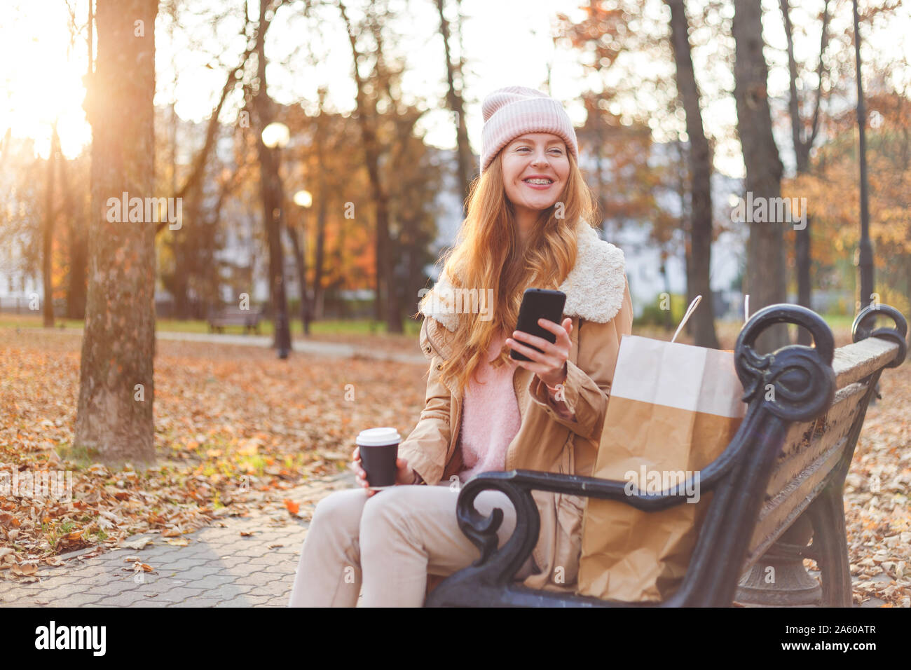 Redhead girl in vestiti caldi seduta con lo smartphone sul banco di legno in autunno park. Foto Stock
