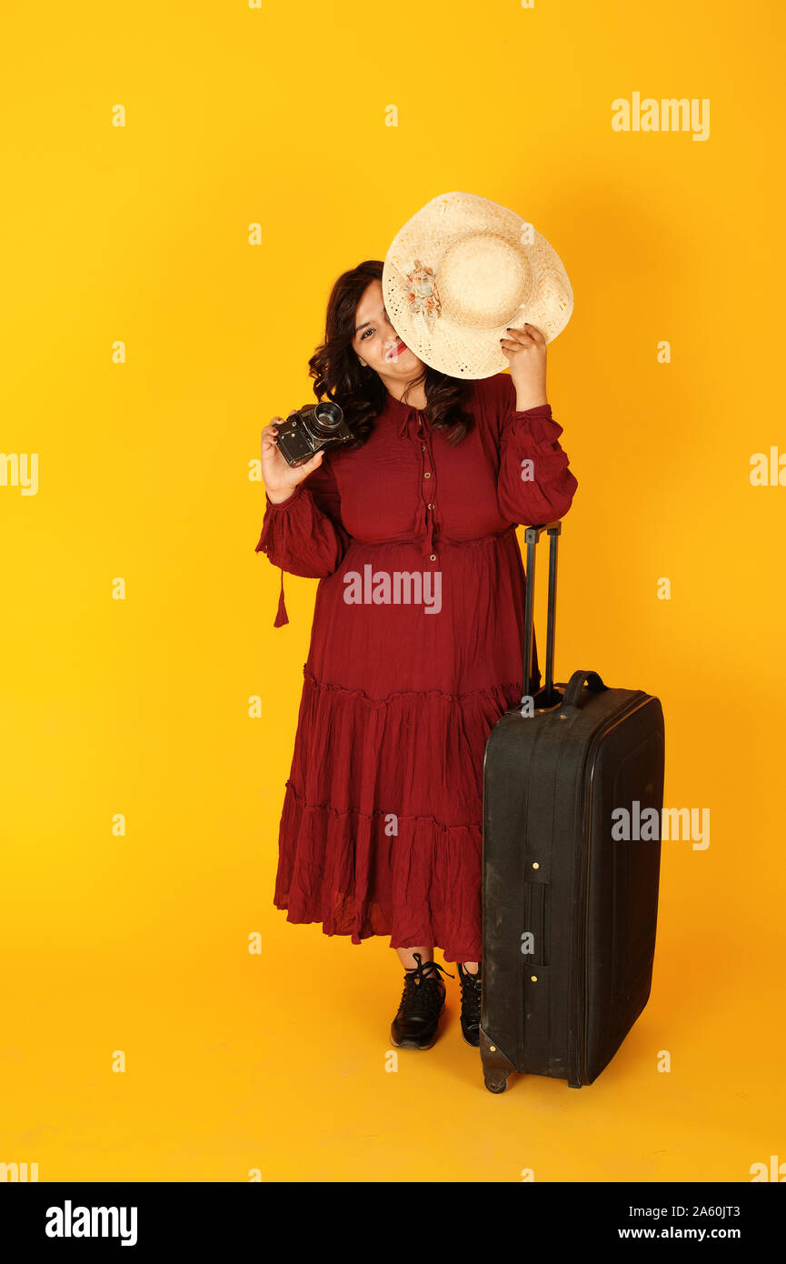 Attraente South Asian traveler donna in rosso profondo abito vestito, hat poste presso lo studio su sfondo giallo con la valigia amd vecchia foto vintage fotocamera. Foto Stock