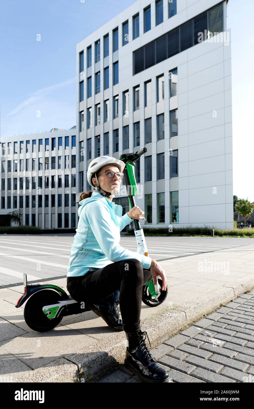 Ritratto di donna matura con lo scooter elettrico seduto sul marciapiede Foto Stock