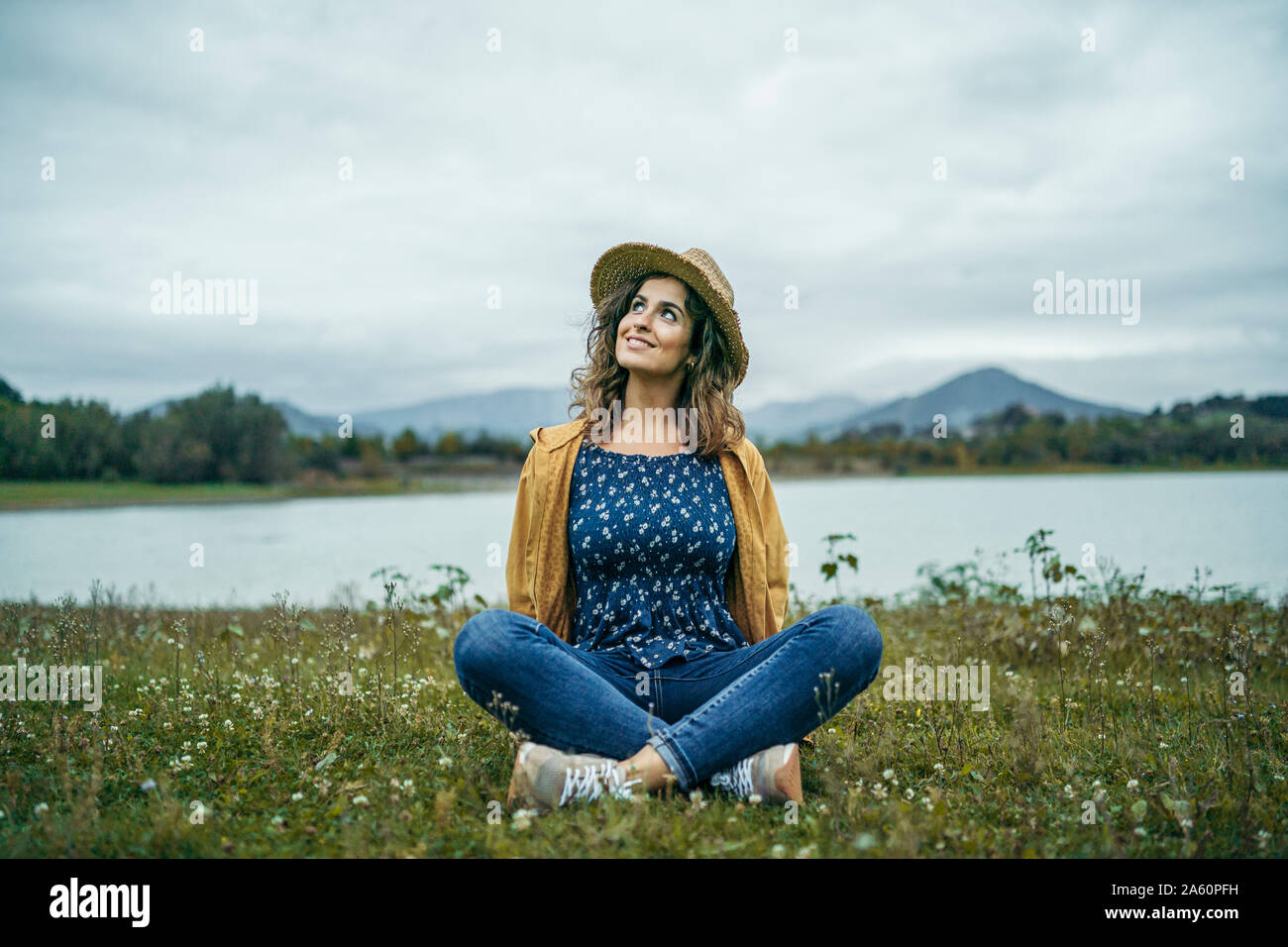 Giovane donna sorridente indossando cappotto giallo seduta su un prato e guardando in alto Foto Stock