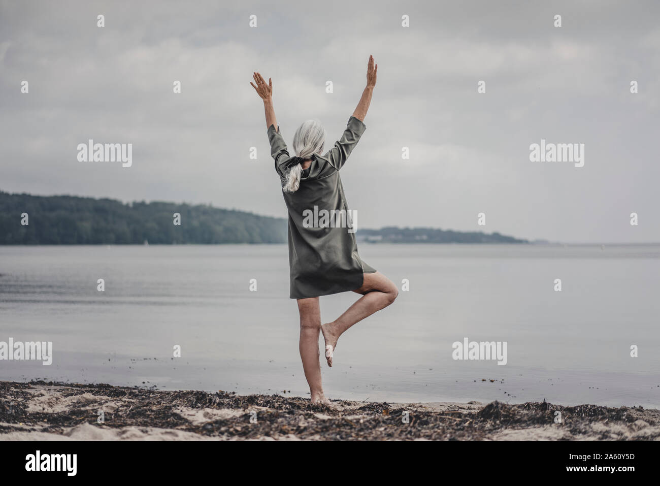 Senior donna che guarda al mare, in piedi su una gamba sola, vista posteriore Foto Stock