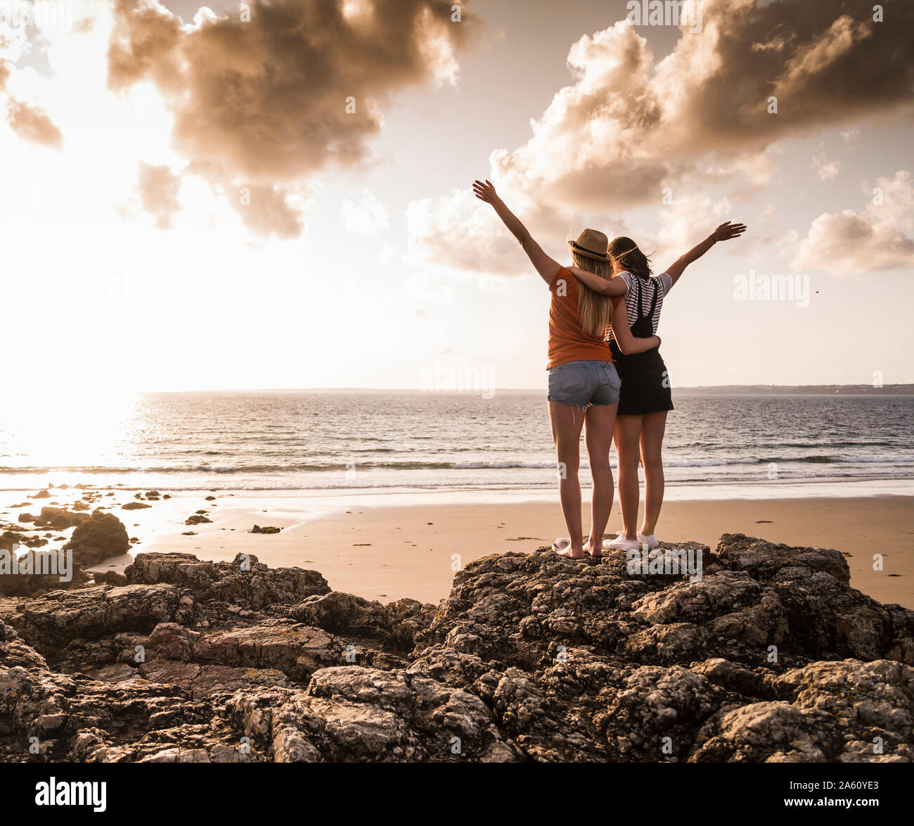 Due amiche permanente sulla spiaggia rocciosa, agitando al tramonto, vista posteriore Foto Stock