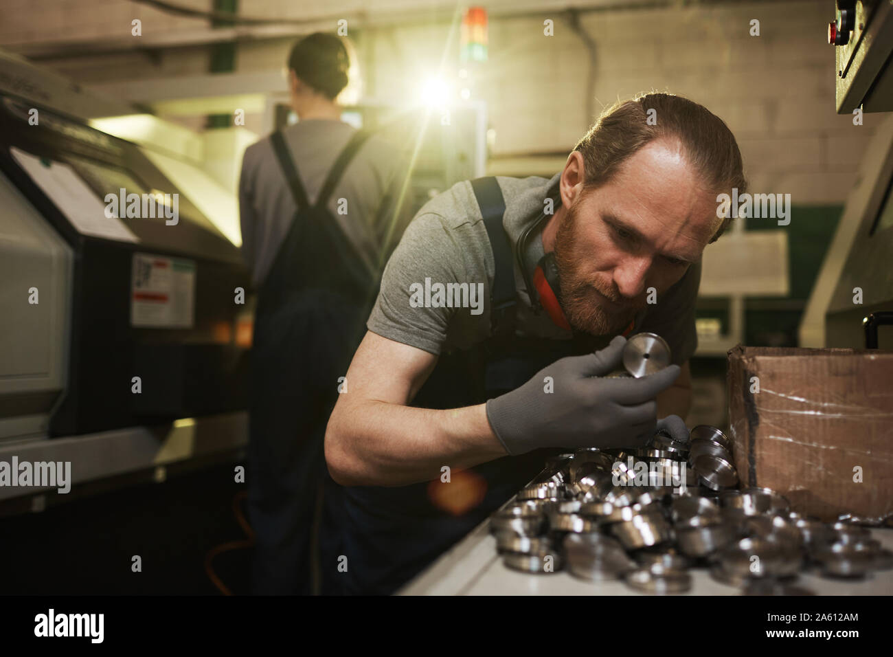 Giovane uomo barbuto in guanti di protezione esaminando i dettagli in metallo mentre si lavora su una fabbrica di metallo con la donna in background Foto Stock