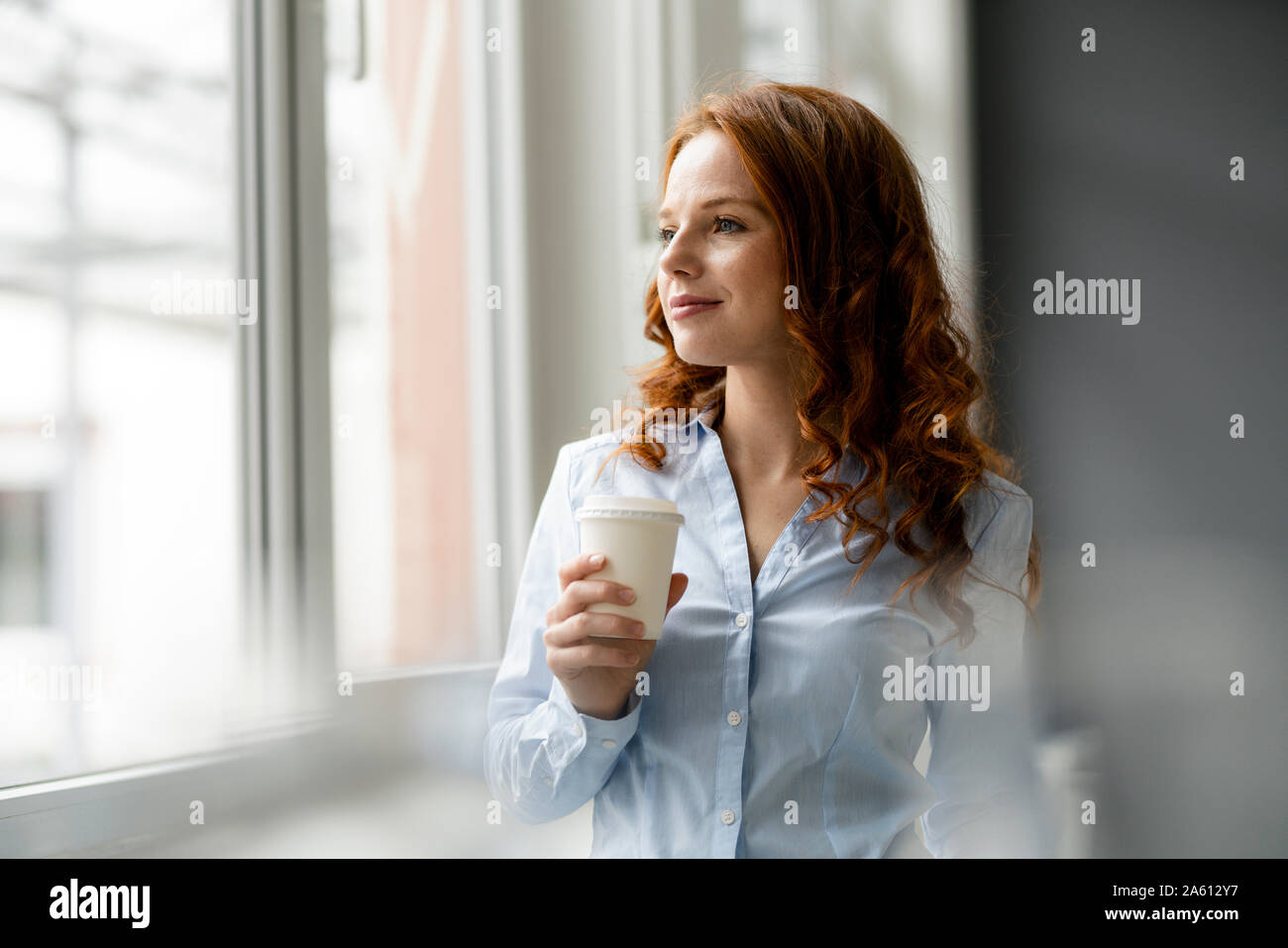 Ritratto di redheaded imprenditrice con caffè per andare guardando fuori della finestra Foto Stock