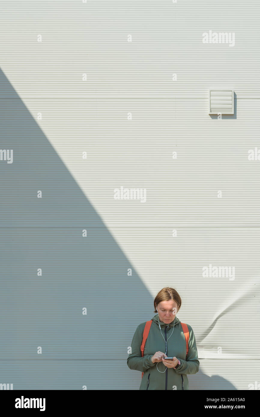Pareggiatore femmina utilizza lo smartphone per la messaggistica di testo sulla strada mentre in piedi accanto alla parete nell'ambiente urbano Foto Stock