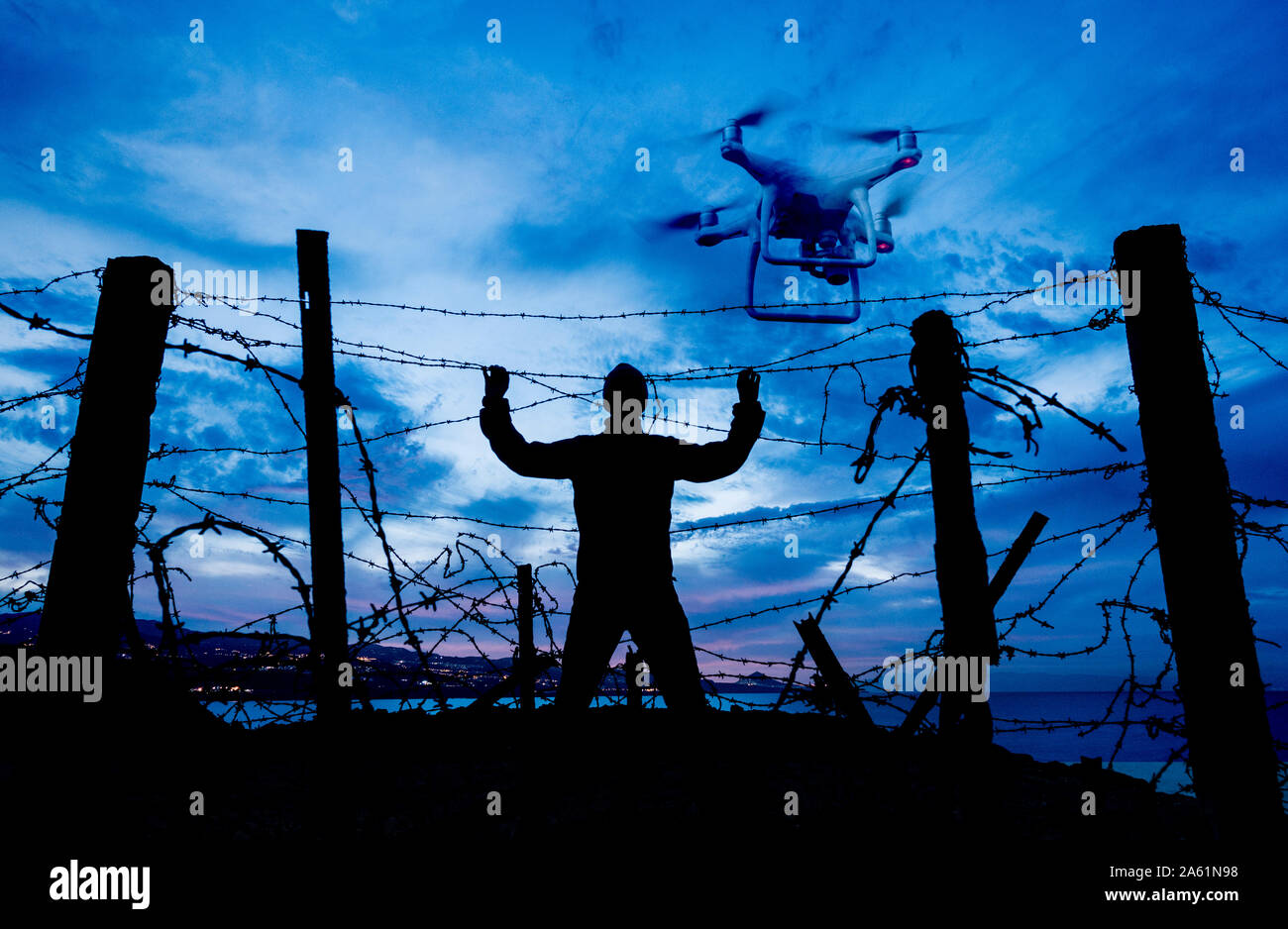 Silhouette di uomo che guarda attraverso recinzione a filo spinato di notte con drone overhead. Brexit, immigrazione, asilo, tecnologia, concetto di controllo delle frontiere. Foto Stock